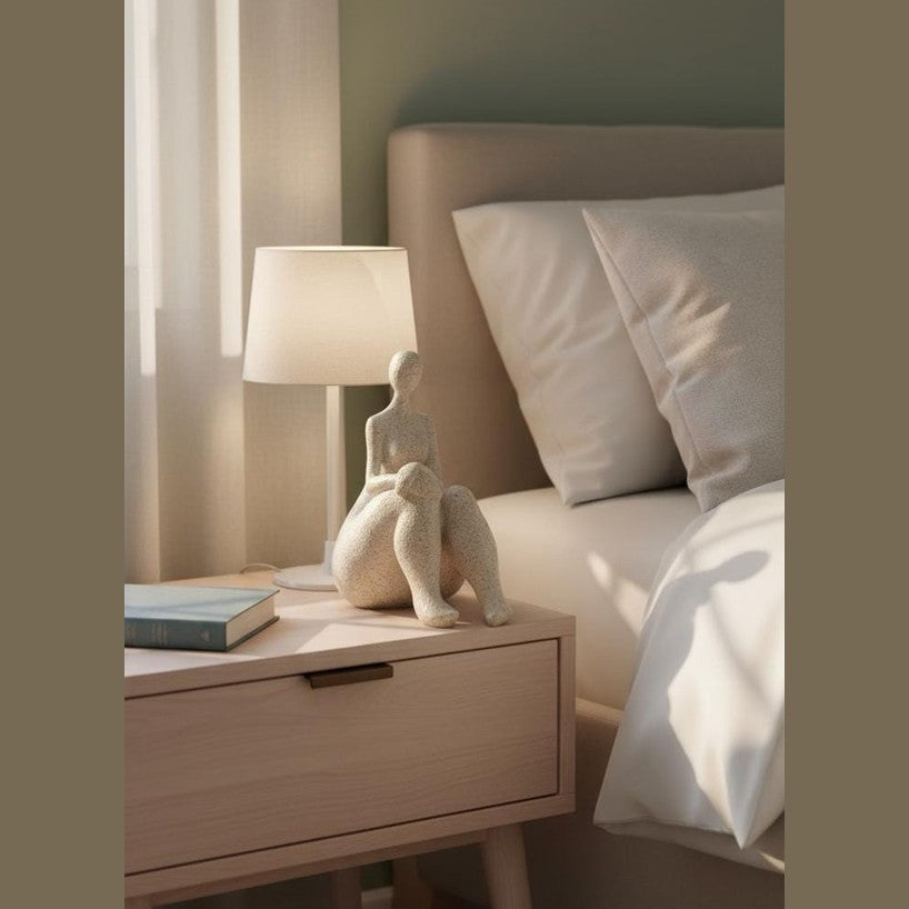 May include: A bedroom scene in neutral tones, featuring a small table with a lamp and a seated figure sculpture. A book rests on the table. A bed with white bedding and pillows is in the background. The room is softly lit.