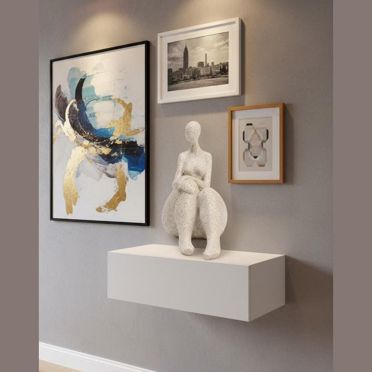 May include: A contemporary wall arrangement with framed art and a sculpture. It features an abstract painting with blue and gold tones, a cityscape photograph, and a geometric print. A white sculpture of a seated figure sits on a white shelf.