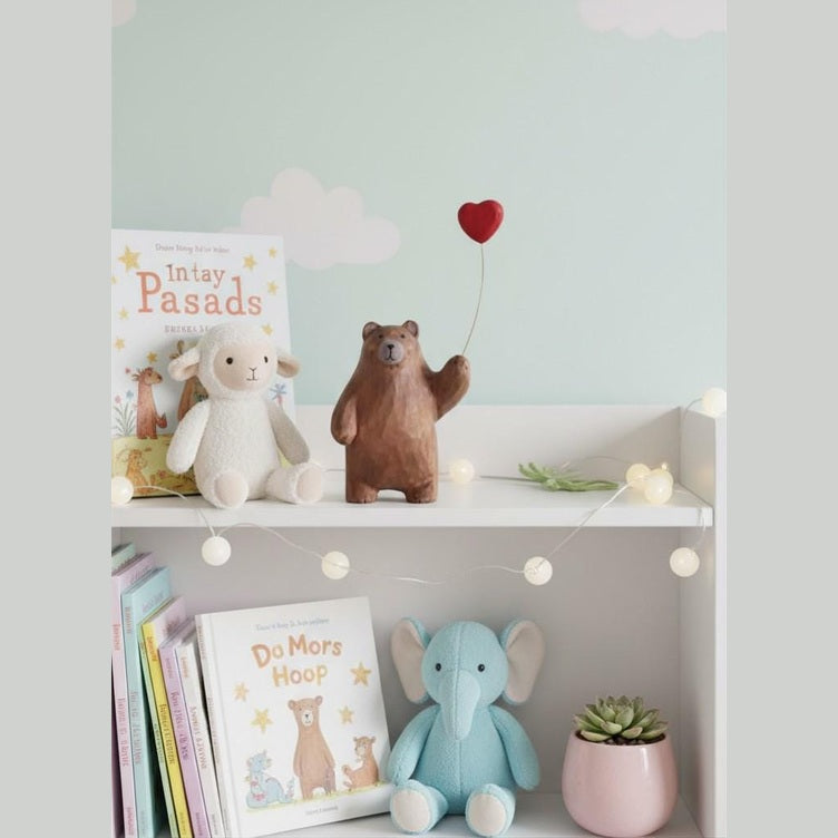 May include: A white shelf in a child's room features a plush lamb, a wooden bear with a red heart balloon, and a blue elephant toy. Books and a pink plant pot are also present, with fairy lights adding a gentle illumination.