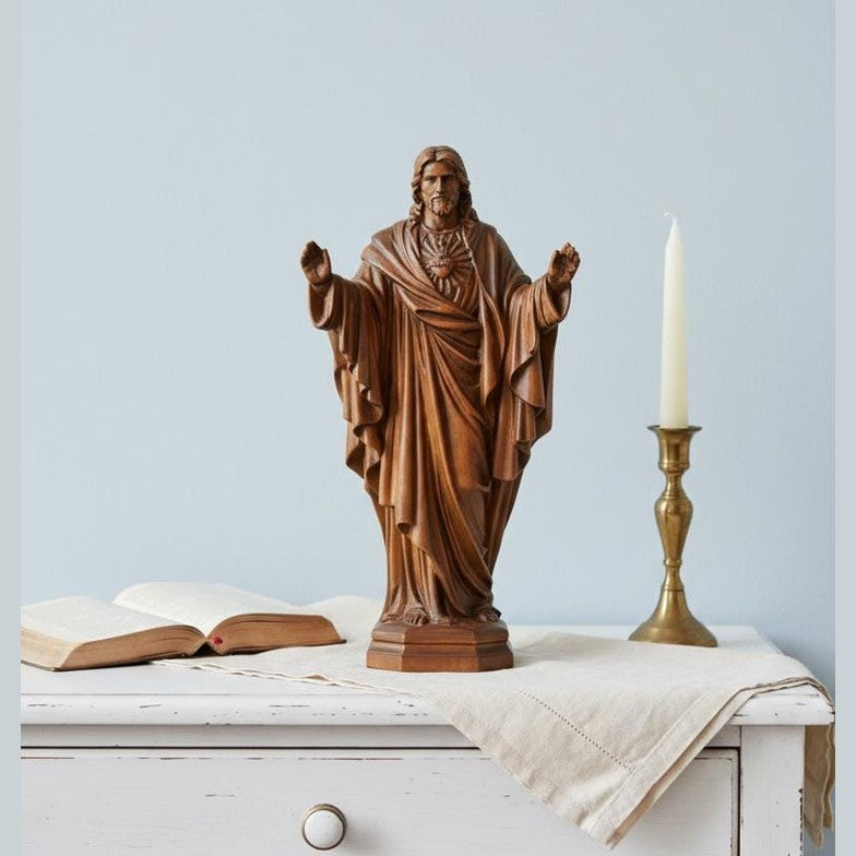 May include: A detailed wooden statue of Jesus with arms outstretched. A lit candle in a brass holder and an open book are on a white table. The statue is brown and stands on a pedestal.